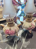 Two antique ceramic table torches with floral patterns and frosted glass chimneys, displayed on a reflective surface.