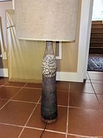 Full view of the tall lamp showing the large beige fabric lampshade and the full height of the heavy textured base standing on a tiled floor.