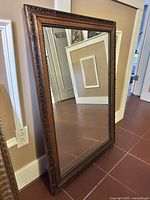 Full view of the large vintage mirror leaning against a wall, showing the reflective glass and entire wooden carved frame.