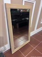 Full view of rectangular mirror leaning against a wall, showing beige colored wood frame and reflective glass surface.