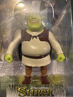 Close-up of Shrek action figure inside clear plastic packaging, showing details of figure's face, clothing, and hands.