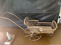 Side front view showing the entire antique wicker doll stroller with curved wooden handles and large spoked metal wheels.