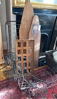 Photo showing two vintage wooden ironing boards, wire laundry basket, wooden tray, and wire cart with wheels arranged upright on floor against a fireplace.