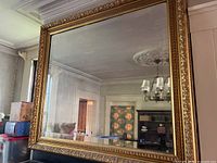 Full view of mirror showing ornate gilt wooden frame and bevelled glass reflecting chandelier and room interior.