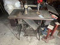 Full view of antique Singer treadle sewing machine mounted in an oak table with the treadle base visible below and surrounding basement items.