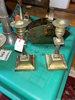 Two brass candle holders with square bases and detailed design on a teal surface