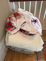 Stack of multiple blankets and comforters showing a red plaid fleece, white sherpa blanket, and light-colored quilts folded on a chair.