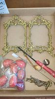 Two ornate brass picture frames (one double hinged) with detailed floral scroll designs, viewed from top with other small items (hand-painted eggs, pins, and a door knocker) around them in a cardboard box.