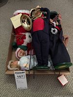 Box containing multiple Byers Choice figurines including the Salvation Army bell ringer and brass instrument player, arranged with other dolls and accessories on a beige carpet.