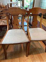 Front view of two wooden chairs with upholstered cream seats, showing their carved backrests and wooden legs.