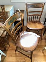 Photo showing two wooden L. Hitchcock chairs with painted panels on the backrest and spindle backs, placed on wood floor near window and other furniture.