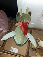 Front view of plush dragon doll showing green head, beige horns, large red tongue, light blue arms, and soft fabric body.