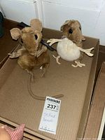 Two plush mice in a cardboard box, the brown one with a long tail and black ribbon, the beige one with a round white belly and purple ribbon.
