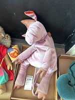 Side view of plush pig doll sitting in a box with a 2"x4" Maxsold sticker for scale, showing long legs, arms, and tail.