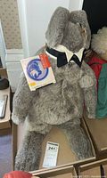 Front side view of the gray plush rabbit with bowtie collar and Forever Toys tag attached, seated on a cardboard box.