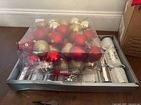 Photo of a box containing Christmas crackers and assorted Christmas baubles in gold, red, silver, and cream colors, metal hooks visible for hanging.