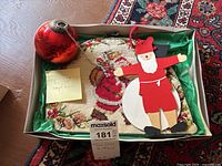 Box showing embroidered tea cozy with stocking design, red ball ornament, and two flat wooden Christmas ornaments (snowman and Santa) on a decorative rug background.