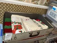 Photo of a suitcase containing folded Christmas-themed linens including a white tablecloth with gift box and Santa Claus print, and other holiday fabrics in red and green.