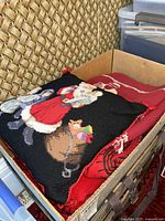 Box containing a folded red holiday table linen and a black needlepoint pillow with Santa Claus and children design.