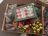 Box containing multiple Christmas ornaments including red and gold baubles, artificial berries, pinecones and other crafting decorative greenery.