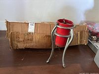 Photo showing the metal Christmas tree stand with red cylindrical holder and silver tripod legs placed in front of the original worn cardboard box for the Christmas tree.