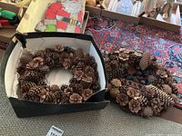 Photo showing two pinecone Christmas wreaths, one inside a black box and the other outside on a patterned rug.