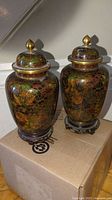 Pair of Japanese cloisonne temple jars with lids, decorated with colorful floral enamel designs and placed on wooden bases.