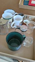 Photo showing varied mugs, creamers, and a glass syrup pitcher arranged in a cardboard box on a table.