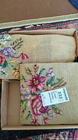 Photo of two unfinished crewel needlepoint projects in a box with floral designs on tan canvas fabric.