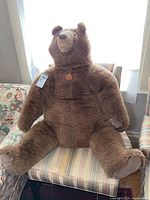 Full view of the large brown teddy bear sitting on a striped chair, showing the entire bear including limbs and face.