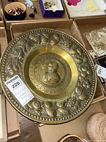 Top-down photo of the brass decorative platter showing the central medallion with historical figure and Latin text, surrounded by embossed fruit and floral border.