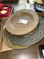 Four brass ornamental wall plates stacked in a box, showing varied metallic tones and embossed designs.