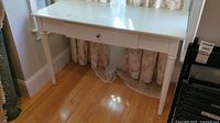 Front and angled view of white painted wooden desk showing the flat top surface, single drawer with knob, and four legs.