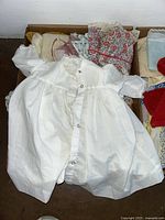 Box with white vintage-style doll dress with lace and buttons on top, along with floral patterned doll dress underneath and other doll clothes visible in the box