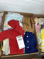 Box with assorted doll clothes including red and blue knit cardigans and light pastel clothes.