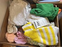 Top view of various doll clothes in a box showing knit sweaters, fabric shoes, and other clothing pieces including green and orange fabrics.