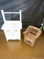 Photo showing white wooden commode/wash stand with towel bar on top and brown woven wicker chair side by side on brown floor.