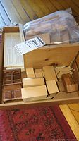 Multiple unfinished wooden dollhouse furniture pieces in natural wood tone in a box with a plastic bag containing smaller pieces and an open instruction book with text and illustrations visible.