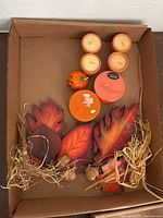 Lot arranged in a cardboard box showing small candles, larger candles, fall leaves, a pumpkin figurine, and raffia filler.