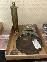 Photo shows a brass cylindrical fireplace match holder with round top and small brass wheels, a tarnished silverplated platter with scalloped edge marked Reed & Barton, an antique metal receipt holder with handle, and a green hexagonal match striker.