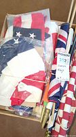 Box containing multiple folded American flags, some with grommets and poles with brass finials arranged vertically in the box