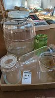 Photo showing five assorted glass jars and storage containers on a cardboard tray on a table, including a large ribbed jar with a white metal lid, and smaller clear and green glass jars with glass lids.