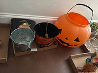Three Halloween treat buckets on a wooden floor. Large orange plastic Jack O Lantern bucket, medium metal pumpkin bucket, and smaller galvanized metal pail with cutout designs. Partial view of Halloween linens and other glass items on the side.