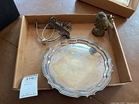 Silver tray with decorative scalloped edges and visible tarnishing inside cardboard box, alongside metal bird figurine and leather strap chain accessory.