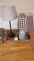 Two table lamps placed side-by-side on the floor in cardboard trays. One lamp features a pottery base and blue checkered shade, the other has a modern metal base and beige shade.