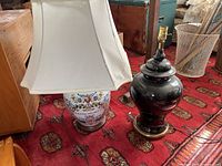 Photo showing two ceramic table lamps: one with a white fabric shade and colorful floral pattern base with 'MASSACHUSETTS' text, and one black ceramic lamp without a shade or harp.