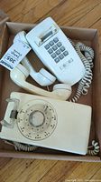 Box containing two vintage landline telephones: one rotary dial phone and one push-button phone, both off-white and showing some discoloration and wear.