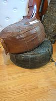 Two round leather footstools stacked, top one brown and the bottom one black, showing wear and seam damage.