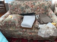 Upholstered loveseat covered with floral and bird pattern slip cover which is visibly ripped, with some pillows and fabric pieces placed on the seat.