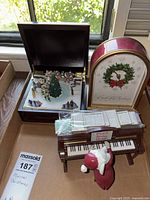 Box containing three mechanical musical Christmas items: a carol clock, a piano with mouse figurine and song cards, a Bavarian ice skating scene music box.